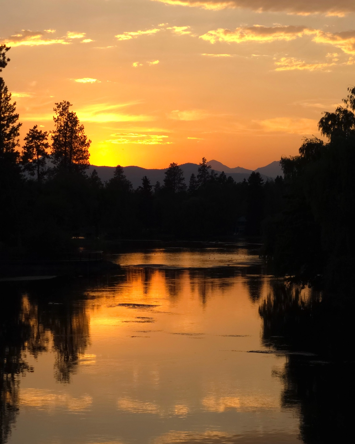 Golden hour on the Deschutes River, Bend Oregon — ponderosa pines reflected in still water with the Cascade silhouette behind